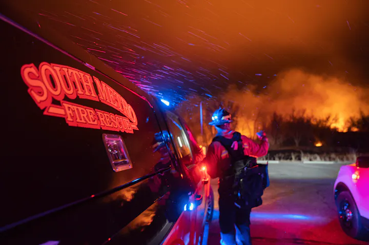 A firefighter next to a vehicle with "South Metro Fire Rescue" on it, emergency lights and a wildfire in the background.