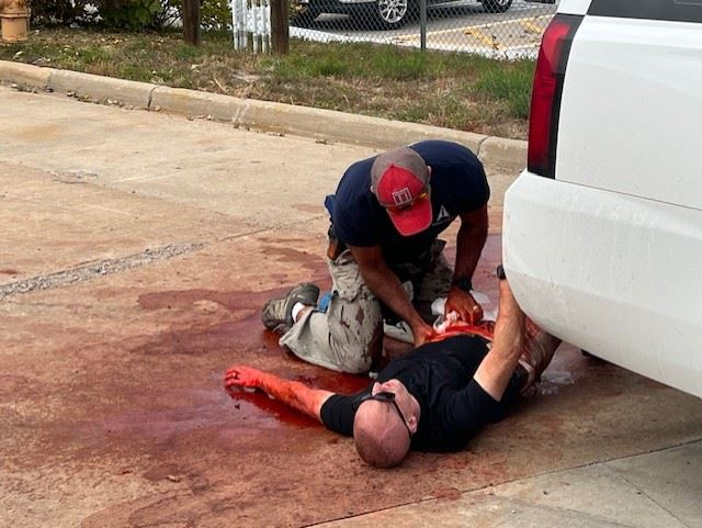 A person aids another lying on the ground, appearing injured with red liquid around them, beside a vehicle.