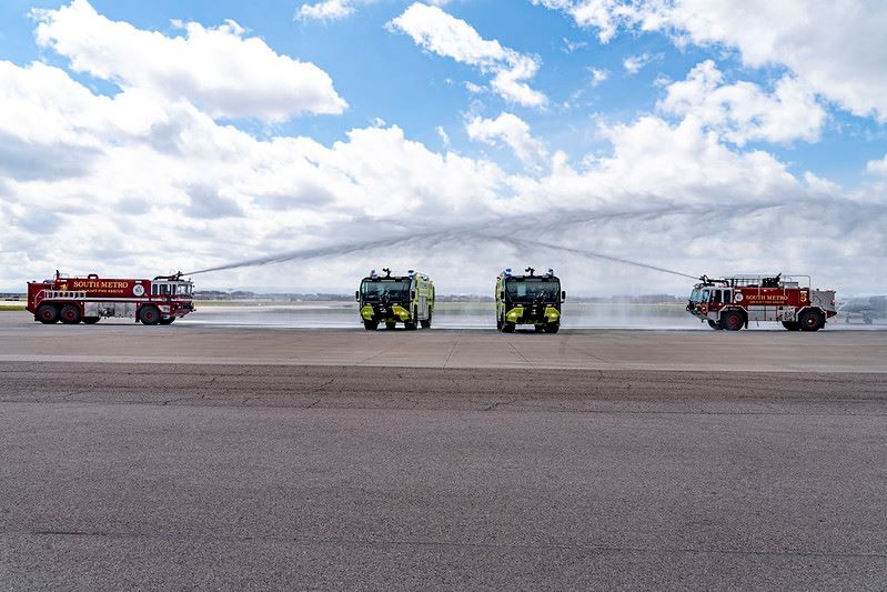 Four fire trucks on a tarmac spraying water arcs against a cloudy sky.