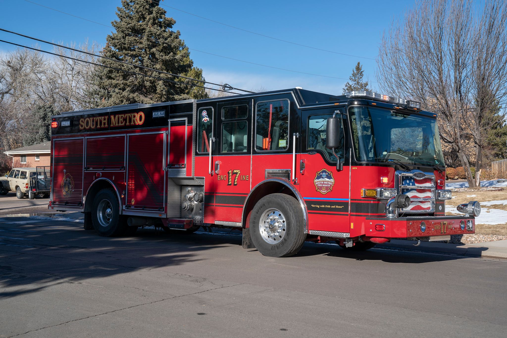 A red and black fire truck labeled "South Metro" is parked on a street with trees and houses in the background.