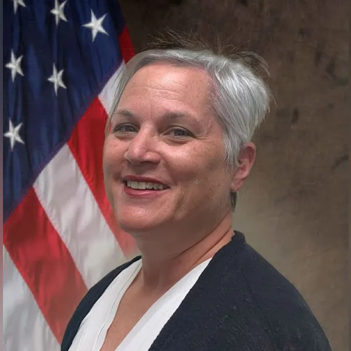 A smiling person with short gray hair in front of a U.S. flag.