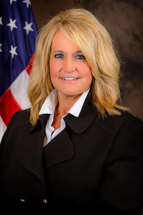 A woman in a black coat with a white collar smiles, standing in front of an American flag.