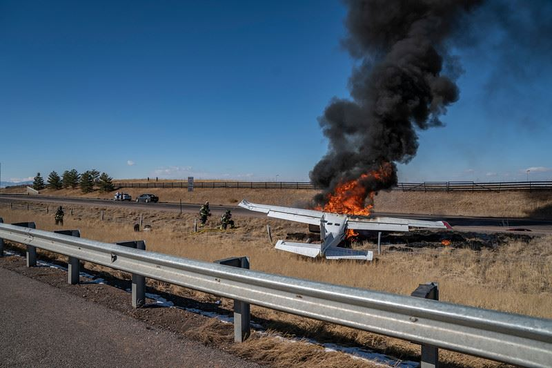 A small plane on fire near a road, with thick black smoke rising. Firefighters and vehicles are nearby.