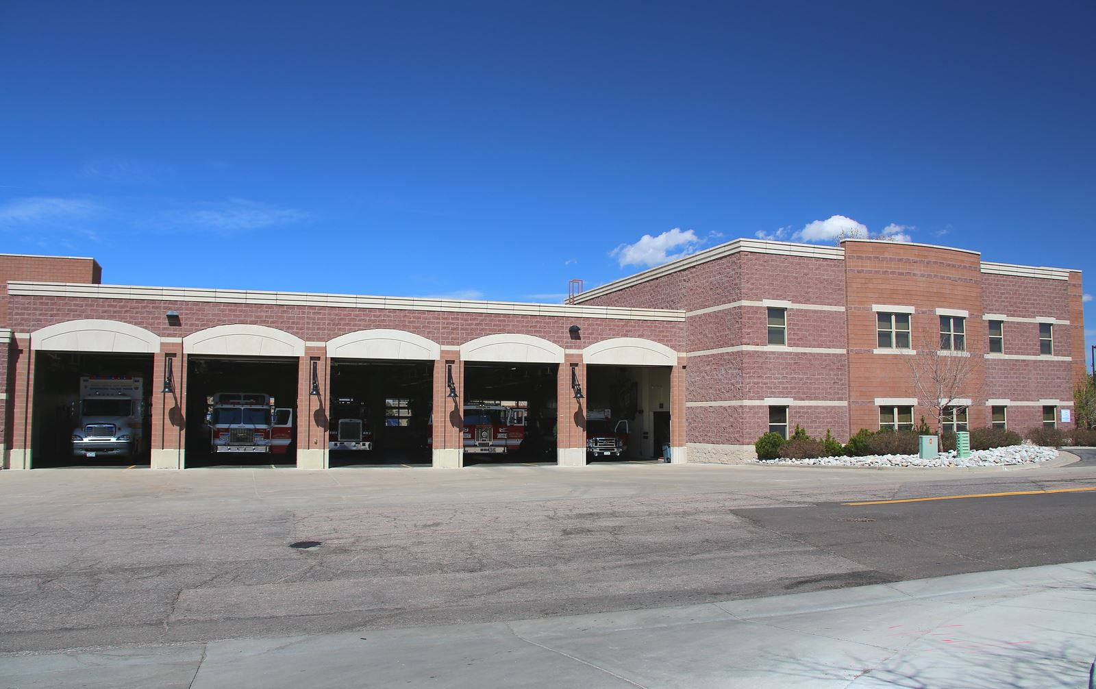 A fire station with parked fire trucks in its open garage bays, set against a clear blue sky.