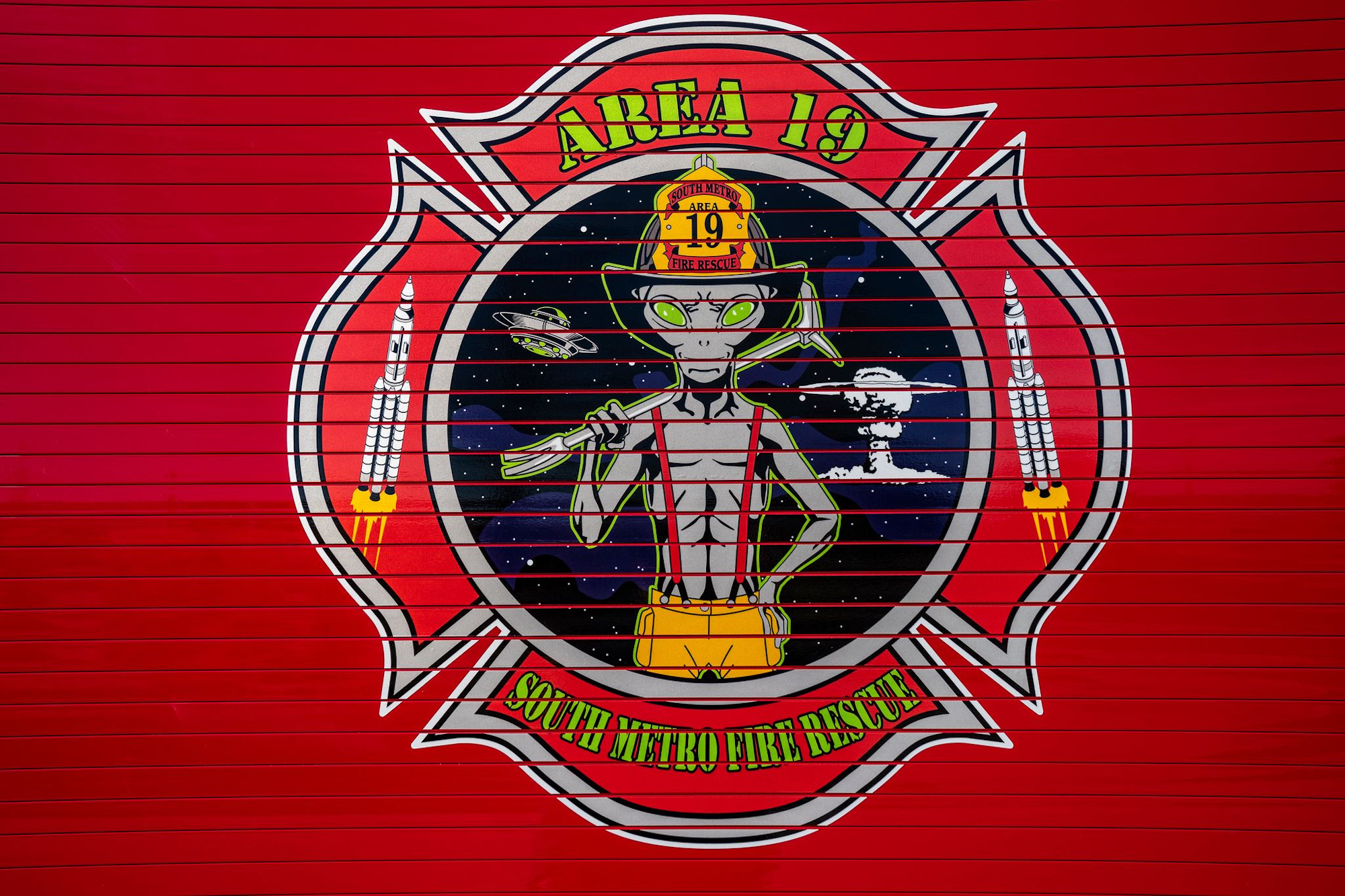 Alien firefighter with "AREA 19" on a fire station door, featuring rockets and UFOs. Text: "South Metro Fire Rescue."