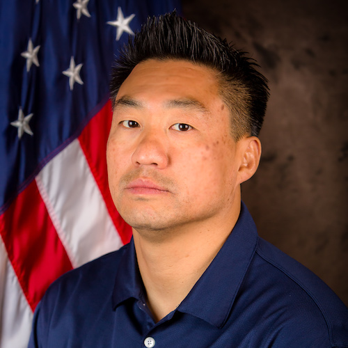 The image features a man in a navy blue shirt, posed in front of a U.S. flag. He has a serious expression.