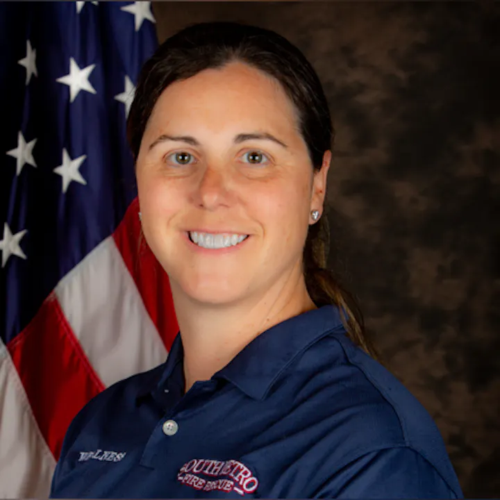 A woman in a blue shirt stands in front of an American flag, smiling at the camera.