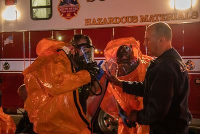 People in orange hazmat suits near a hazardous materials vehicle; one person holding a device.
