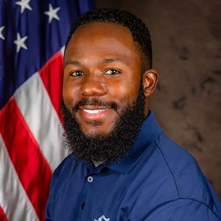 A smiling man with a beard, wearing a blue shirt, poses in front of an American flag.