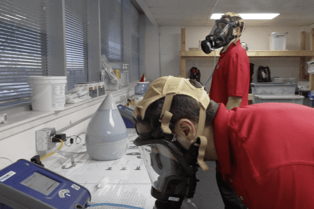 Two people in red shirts wear gas masks and perform lab work, using electronic equipment in an indoor lab setting.
