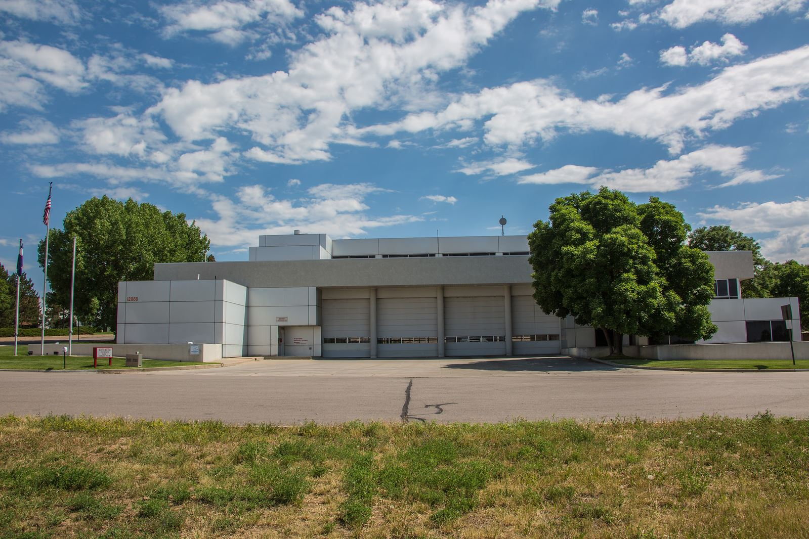 A large, modern building with garage doors, surrounded by trees and flags, under a blue sky with clouds.