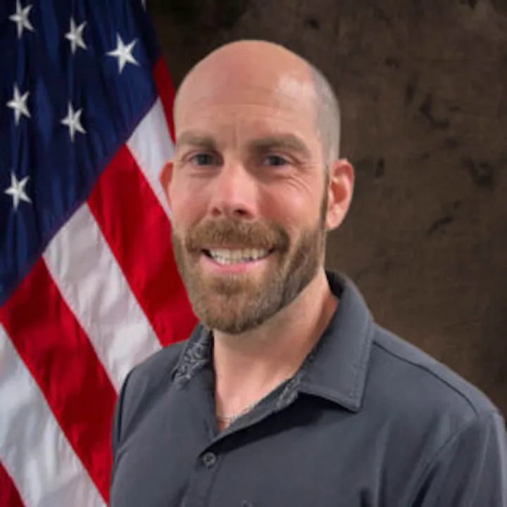 The image shows a smiling man in a dark shirt, standing in front of an American flag.