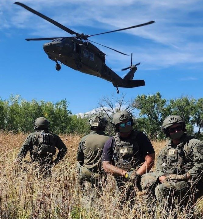 Soldiers in gear sit in a field as a helicopter flies overhead.