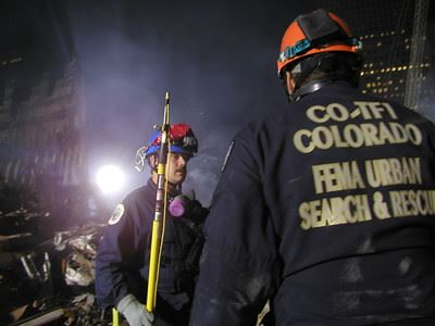 Two rescue workers in protective gear at a disaster site, with debris and construction tools visible.