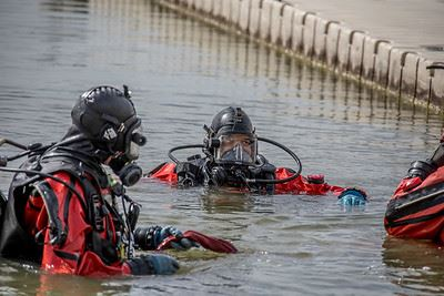 Divers in red dry suits and black helmets in the water near a dock.