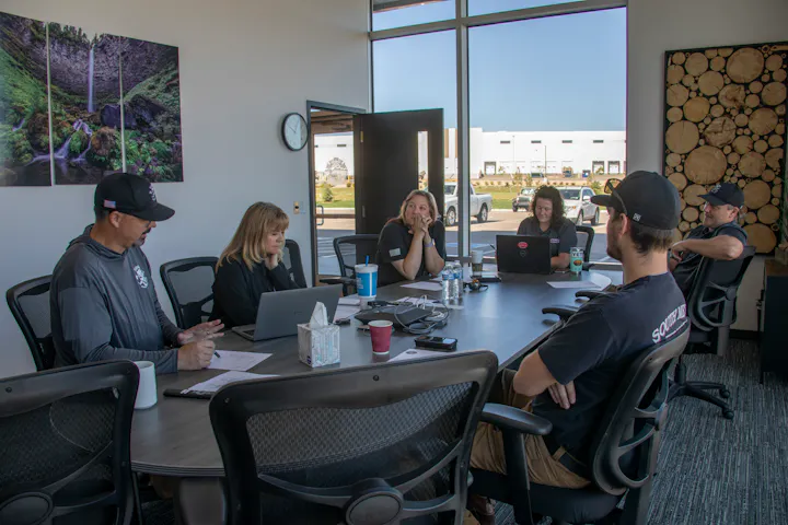 A group of six people is engaged in a meeting around a table, using laptops and discussing in a well-lit office space.