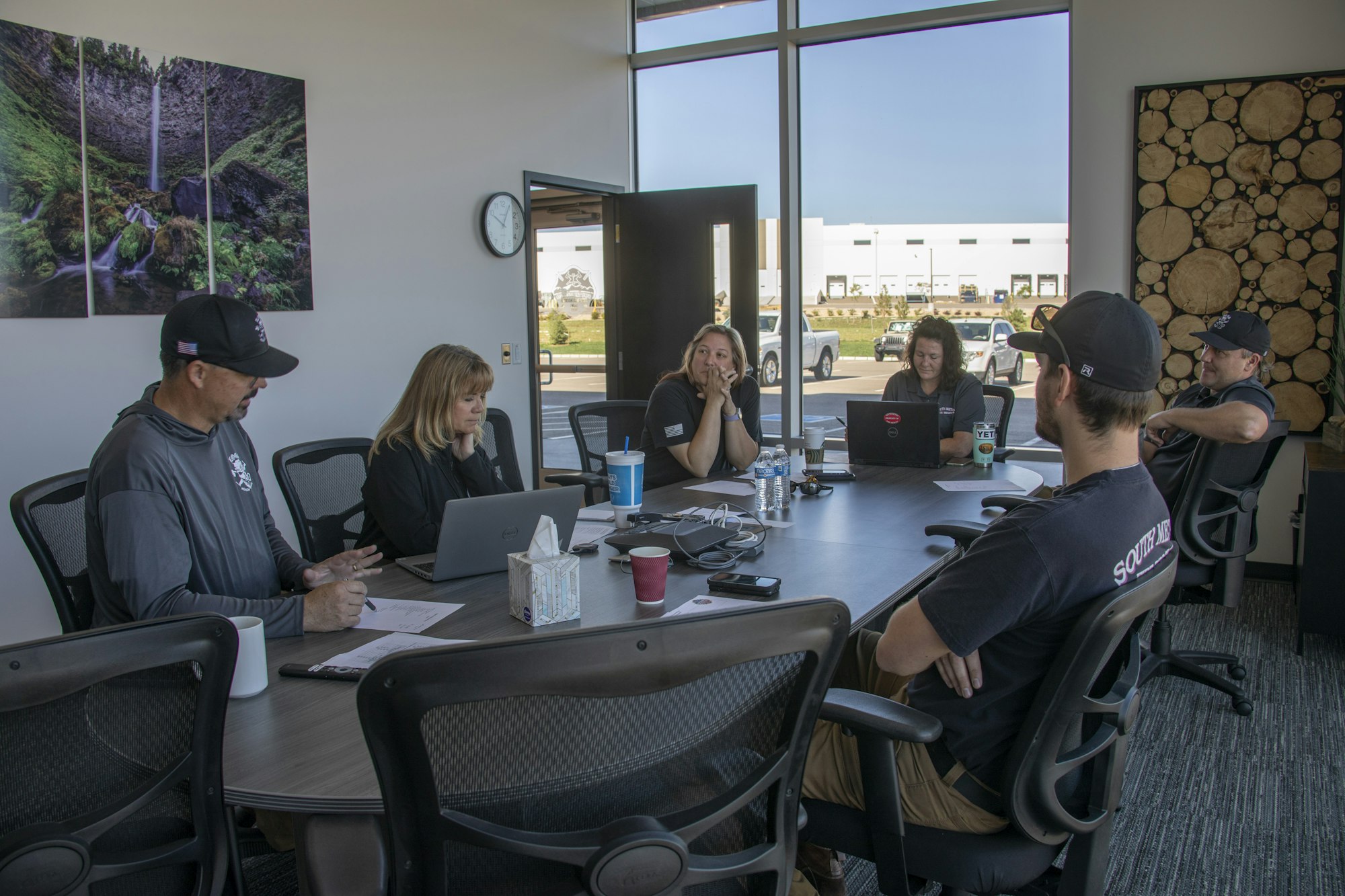 A group of six people is engaged in a meeting around a table, using laptops and discussing in a well-lit office space.