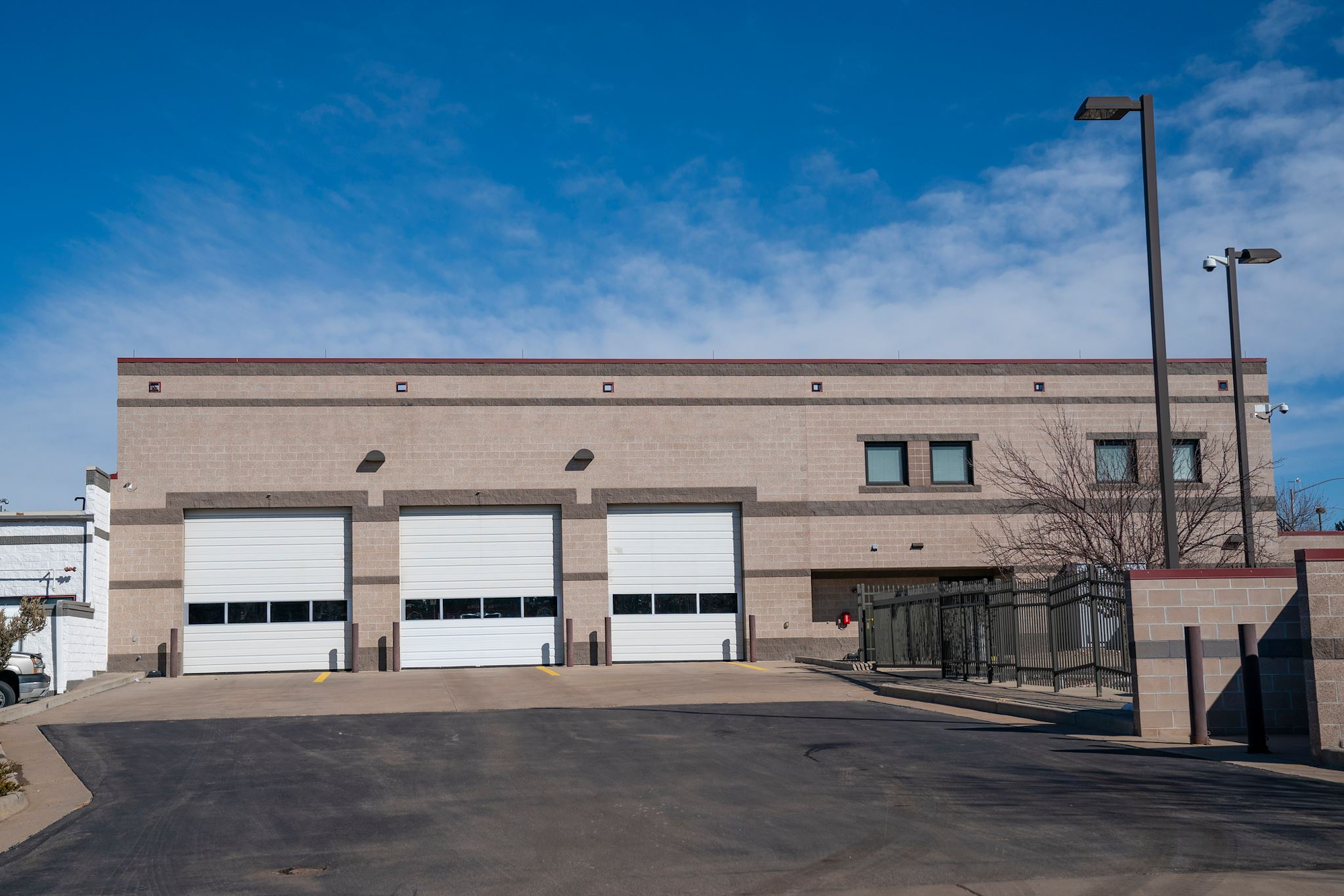 A beige industrial building with three large garage doors, two windows, and surrounding streetlights under a clear blue sky.