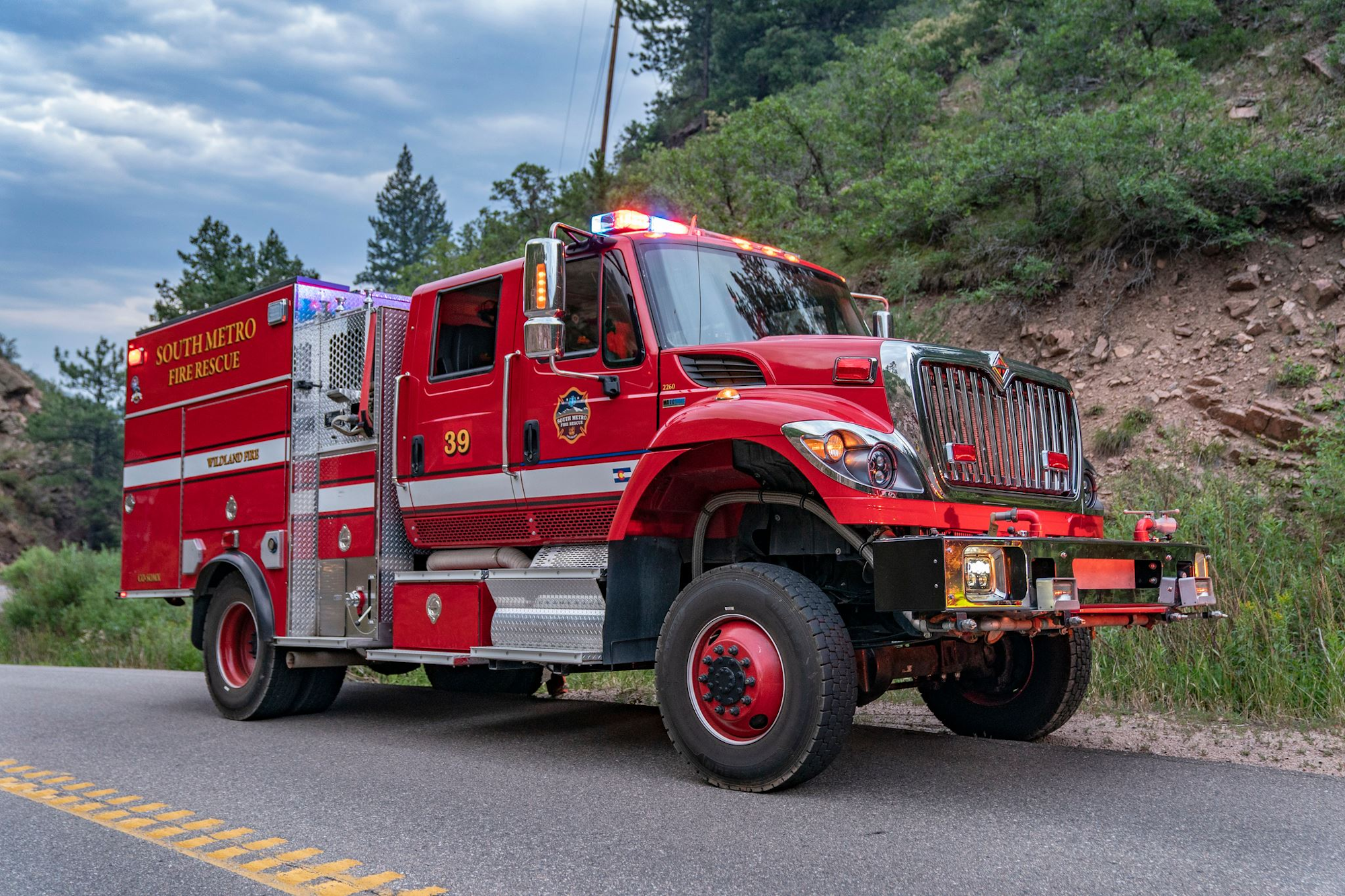 Red fire rescue truck with lights on, parked on a road in a forested area.