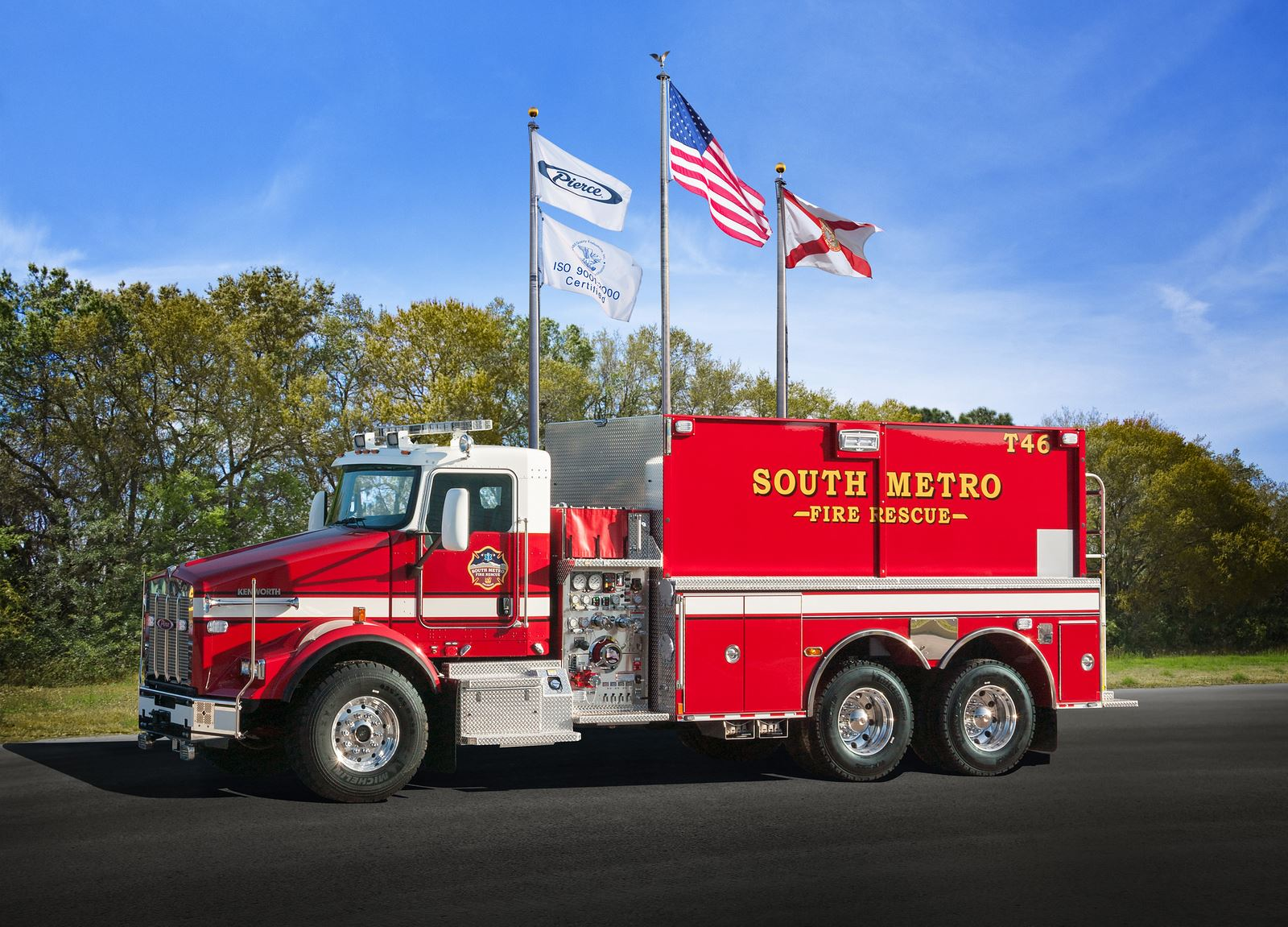 Red fire rescue truck parked, marked "South Metro," with three flags flying above, against a backdrop of trees and blue sky.