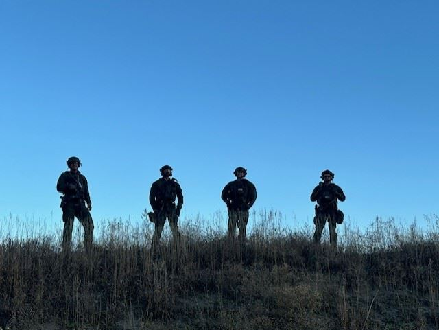 Four people in tactical gear standing in a line on a grassy hill, silhouetted against a clear blue sky.