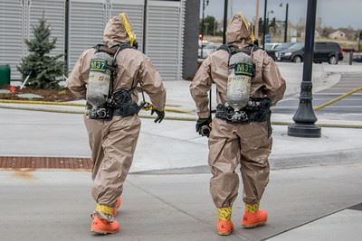 Two people in hazmat suits with backpacks and masks walk down a street.