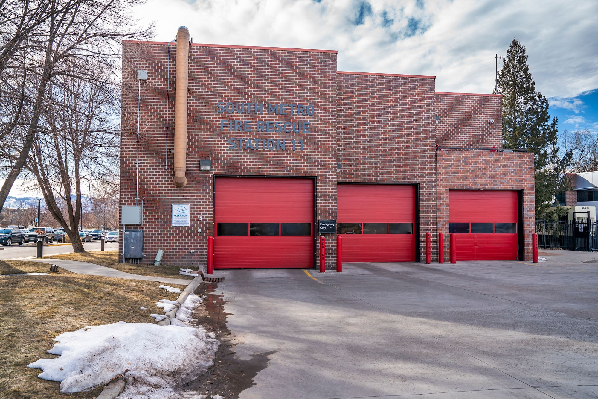 A fire station with three red garage doors and brick exterior. Snow and trees are in the foreground.