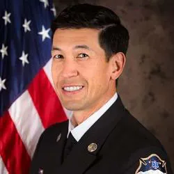 A person in a formal uniform stands in front of an American flag.
