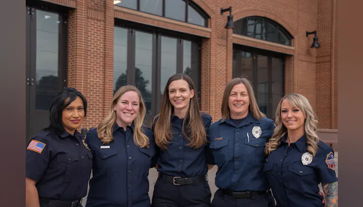 Five women in uniform, likely emergency responders, standing in front of a building with large windows.