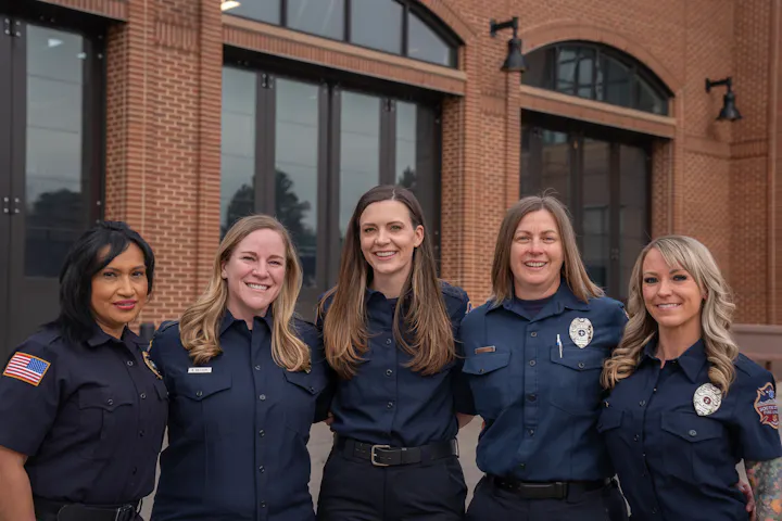 Five women in uniform, likely emergency responders, standing in front of a building with large windows.