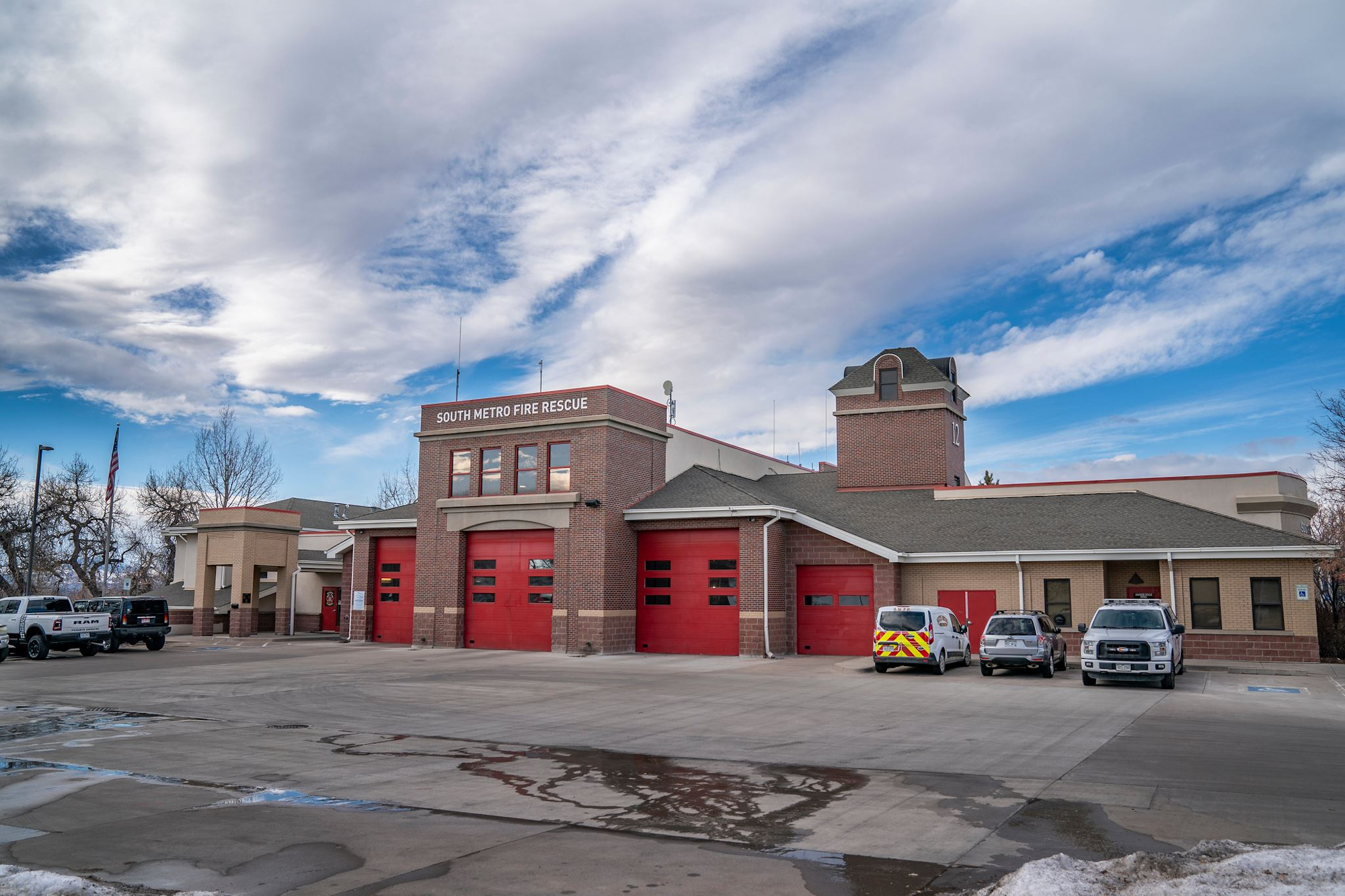 A fire station with red doors, vehicles parked in front, and an American flag on a cloudy day.