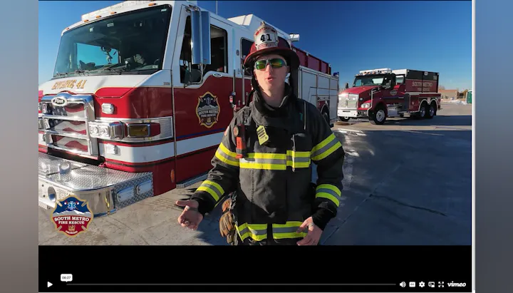 A firefighter in gear stands in front of fire trucks, likely discussing fire safety or rescue operations.