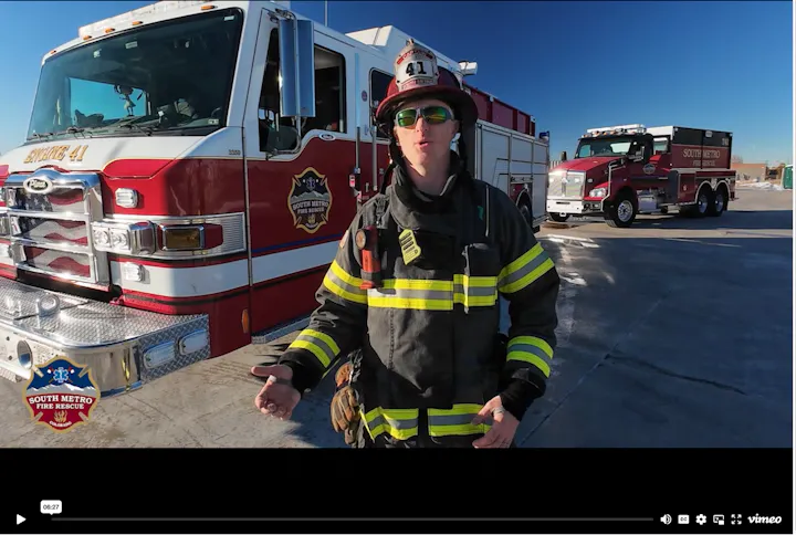 A firefighter in gear stands in front of fire trucks, likely discussing fire safety or rescue operations.