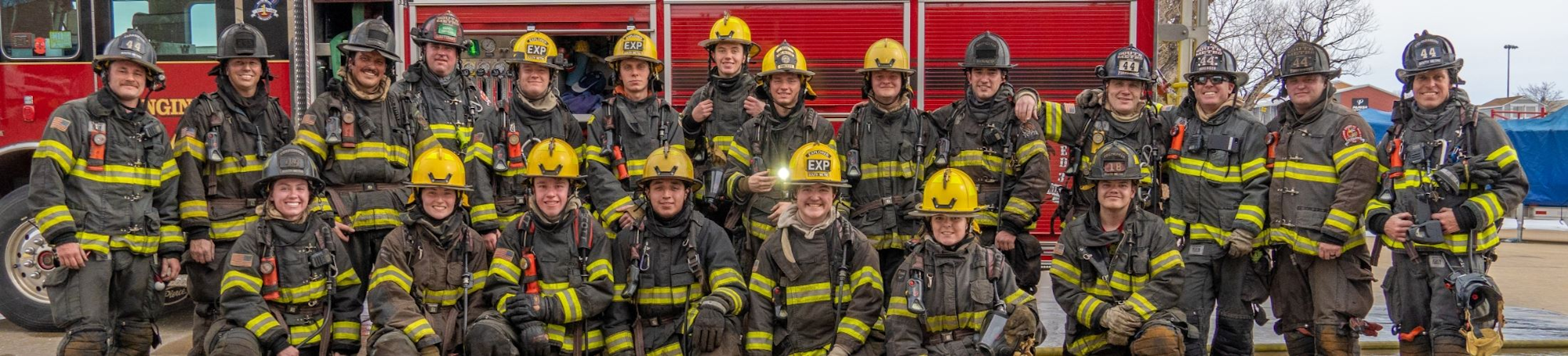 A group of firefighters posing in front of a fire truck, wearing helmets and uniforms.