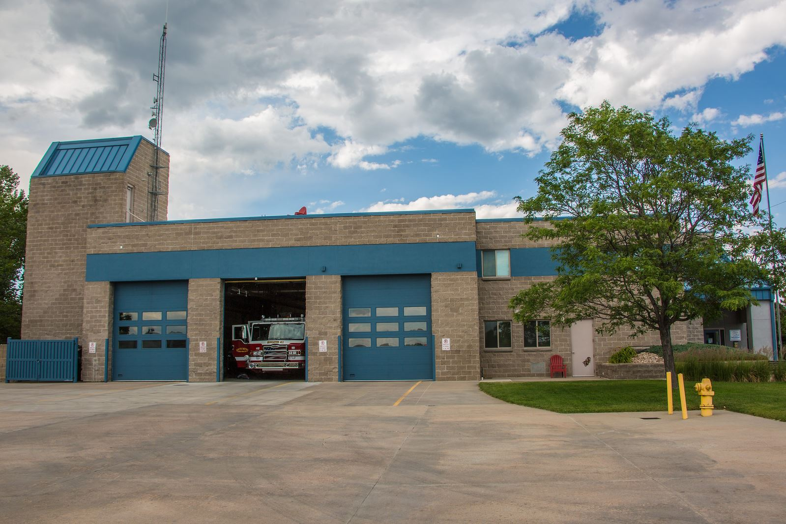 Fire station with parked fire truck, blue garage doors, tree, flag, and cloudy sky.