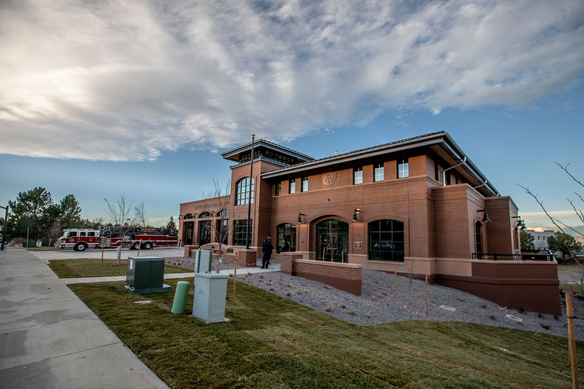 A fire station with a red fire truck parked outside, surrounded by a grassy area and a cloudy sky above.