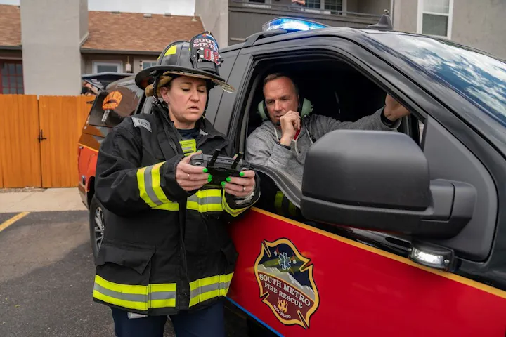 A firefighter operates a remote control next to a "South Metro Fire Rescue" vehicle with a man seated inside.