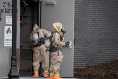 Two people in hazmat suits and masks enter a building through a door labeled with warning signs.