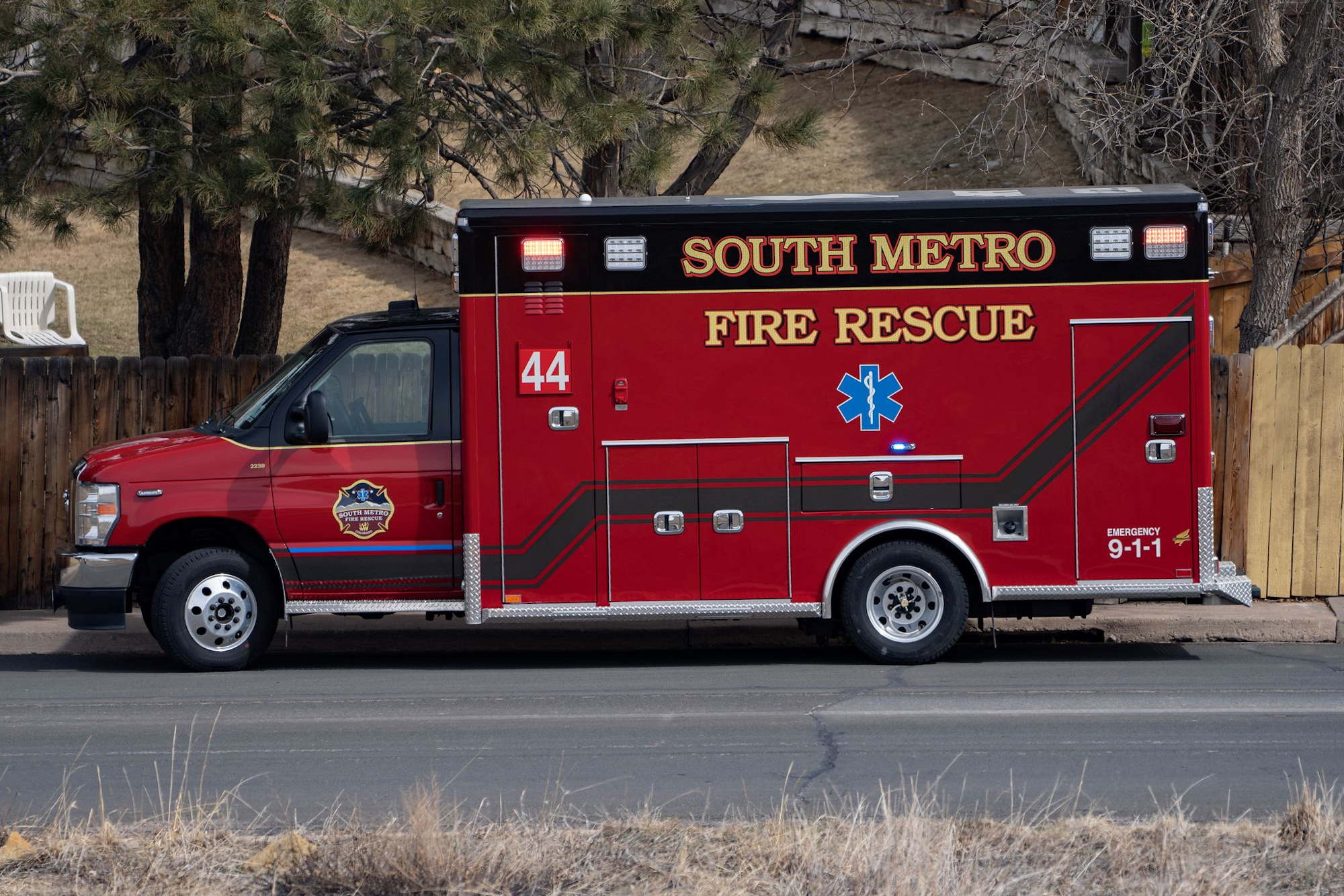 A red South Metro Fire Rescue ambulance parked on a street next to a fence and trees.