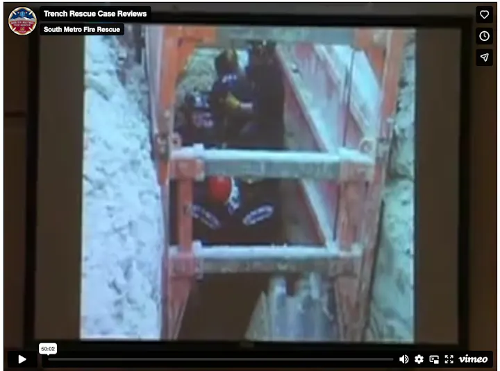 The image shows a trench rescue operation, with rescue workers assisting in a confined space surrounded by protective barriers.