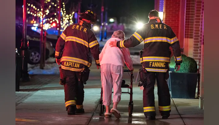 Two firefighters assisting an elderly person with a walker on a city sidewalk.