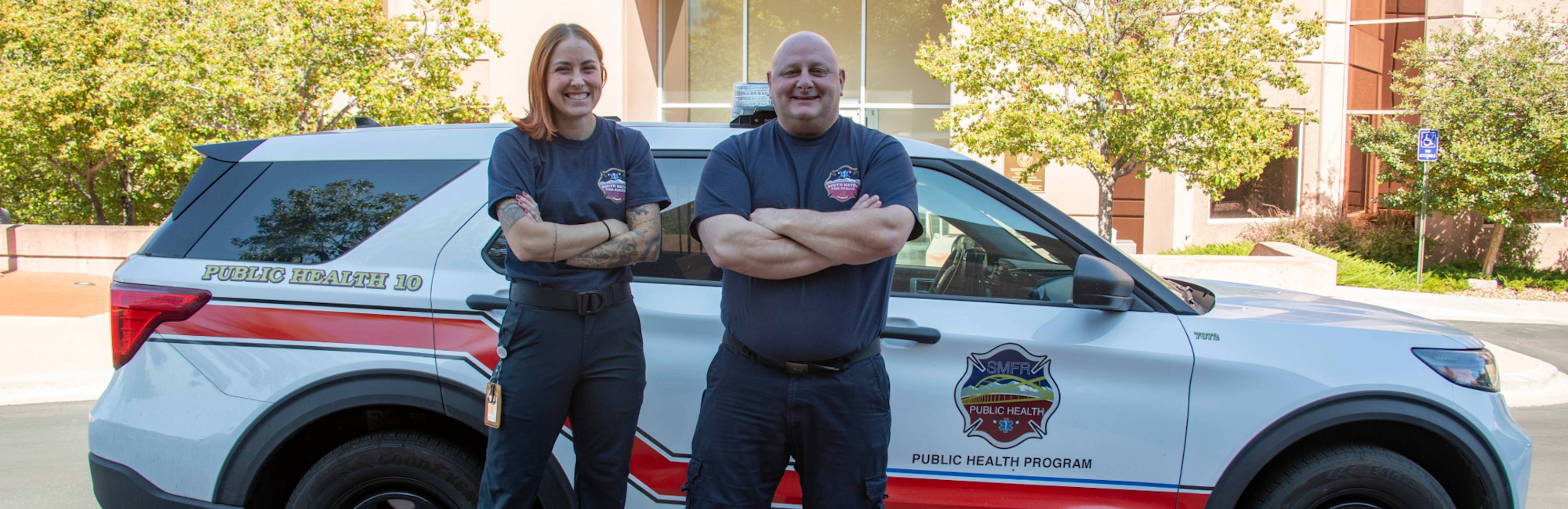 Two people smiling, standing in front of a "Public Health Program" vehicle.