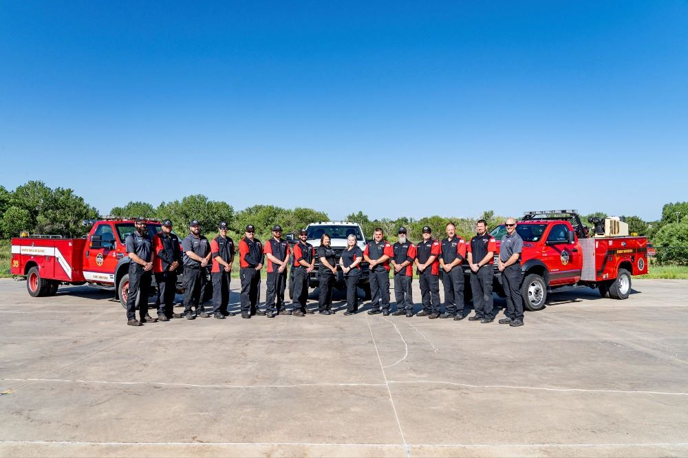 A group of firefighters stands in front of red fire trucks outdoors on a sunny day.