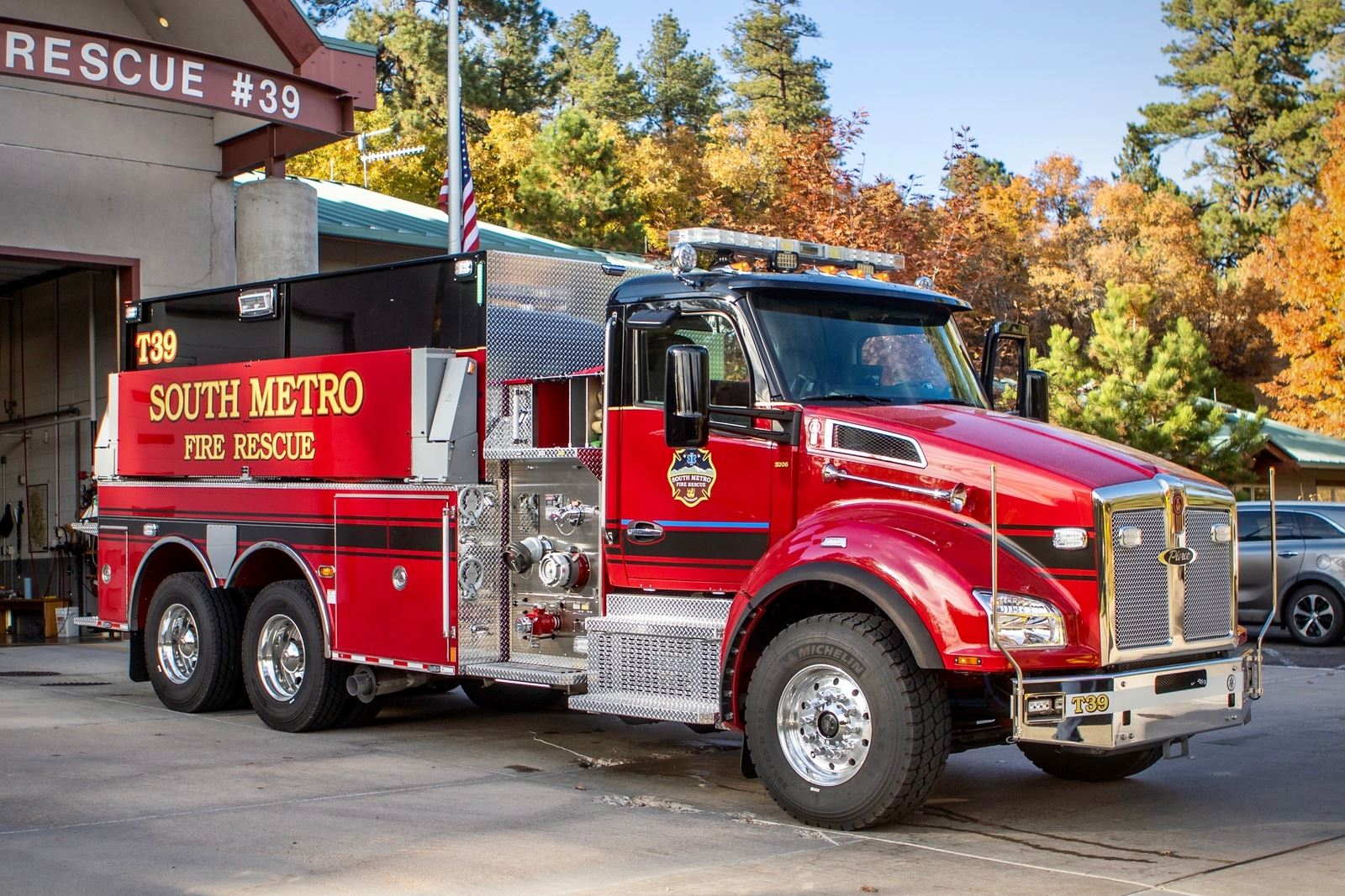 Red fire rescue truck labeled "SOUTH METRO" parked outside a fire station with autumn trees in the background.