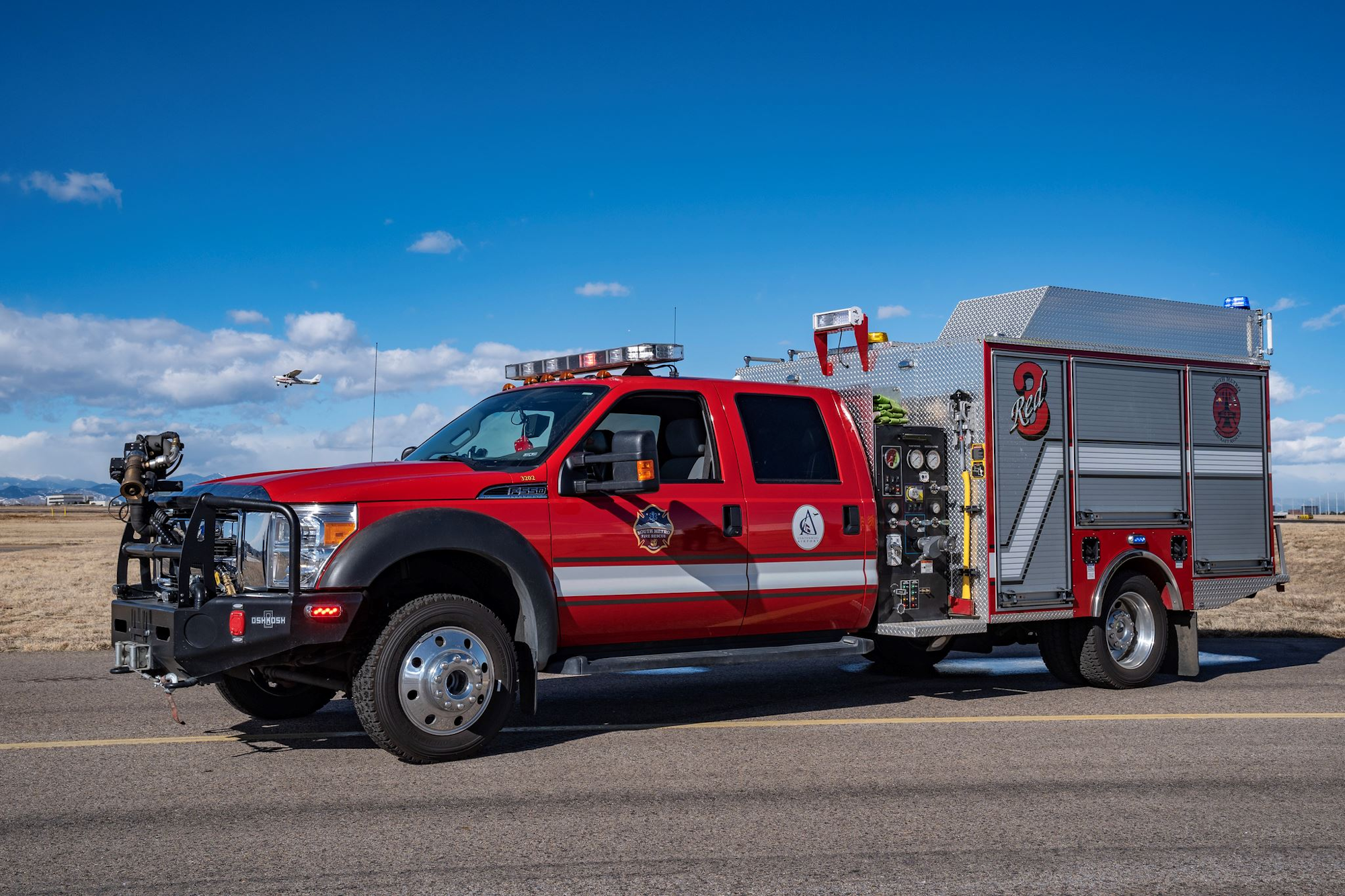 Red airport fire truck on a runway, with a small plane in the sky above a grassy field.