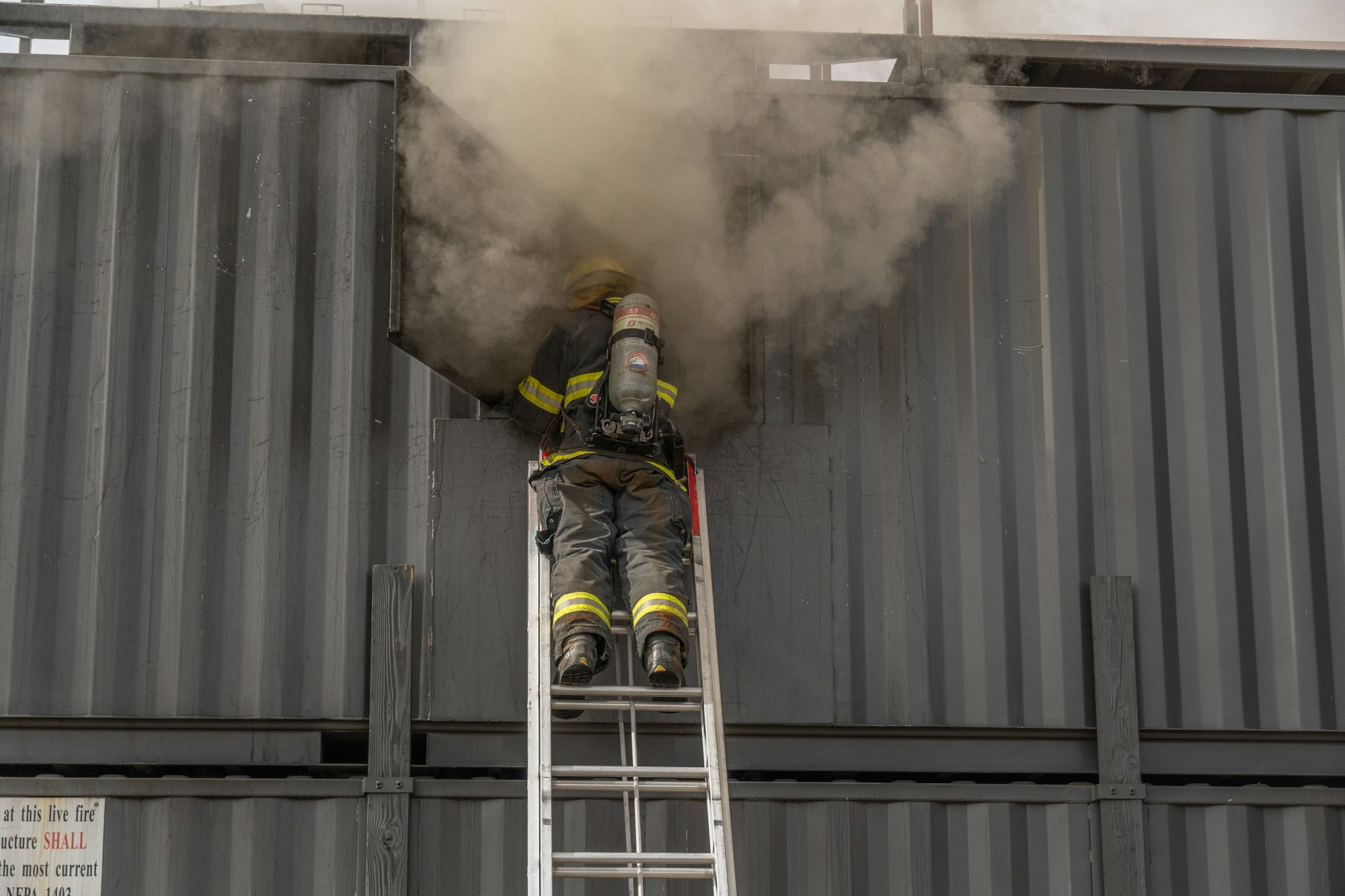 A firefighter climbs a ladder to address smoke emerging from an opening in a structure, likely during a training exercise.
