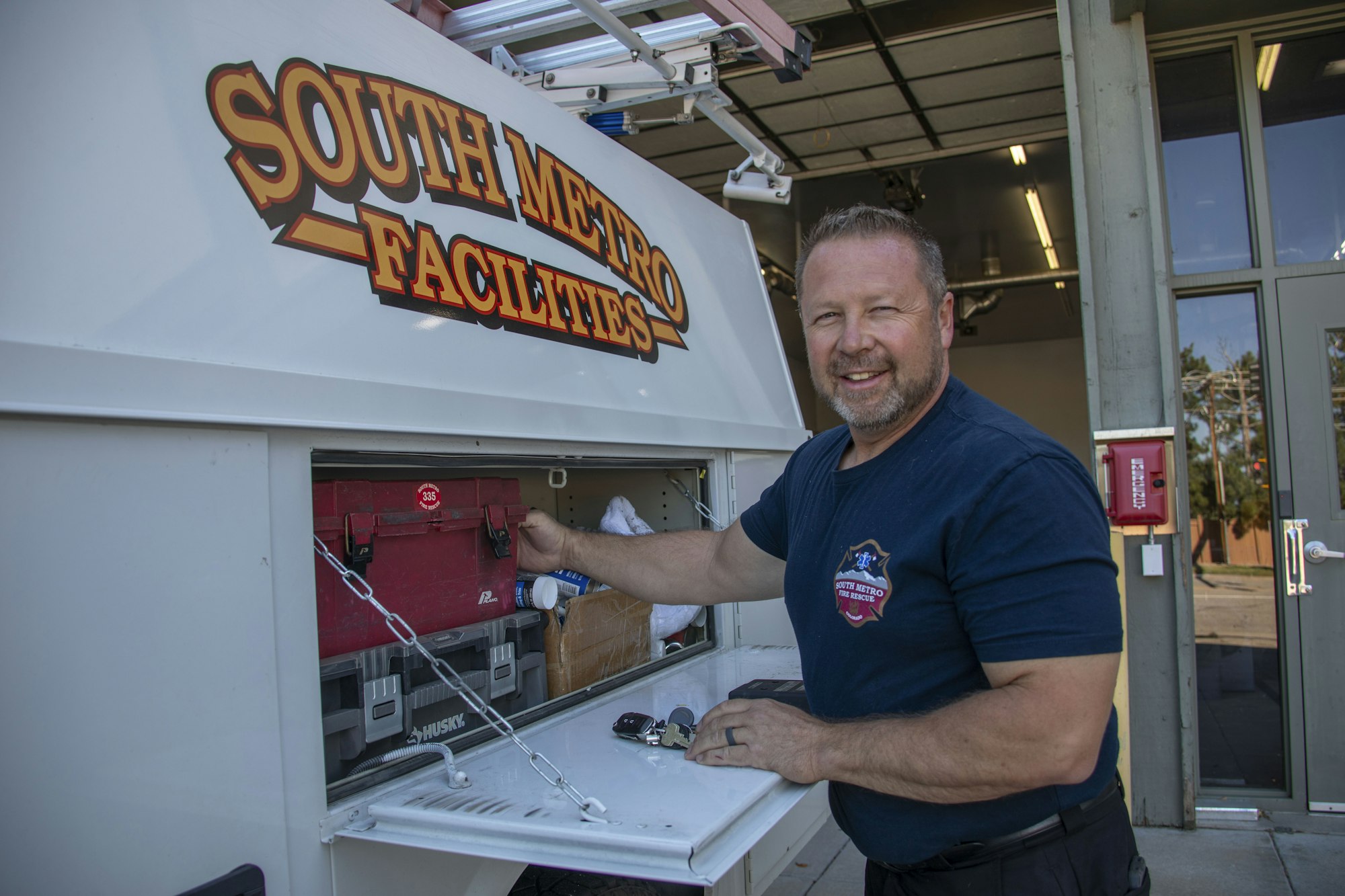 A man is smiling while organizing tools in a service vehicle marked "South Metro Facilities."