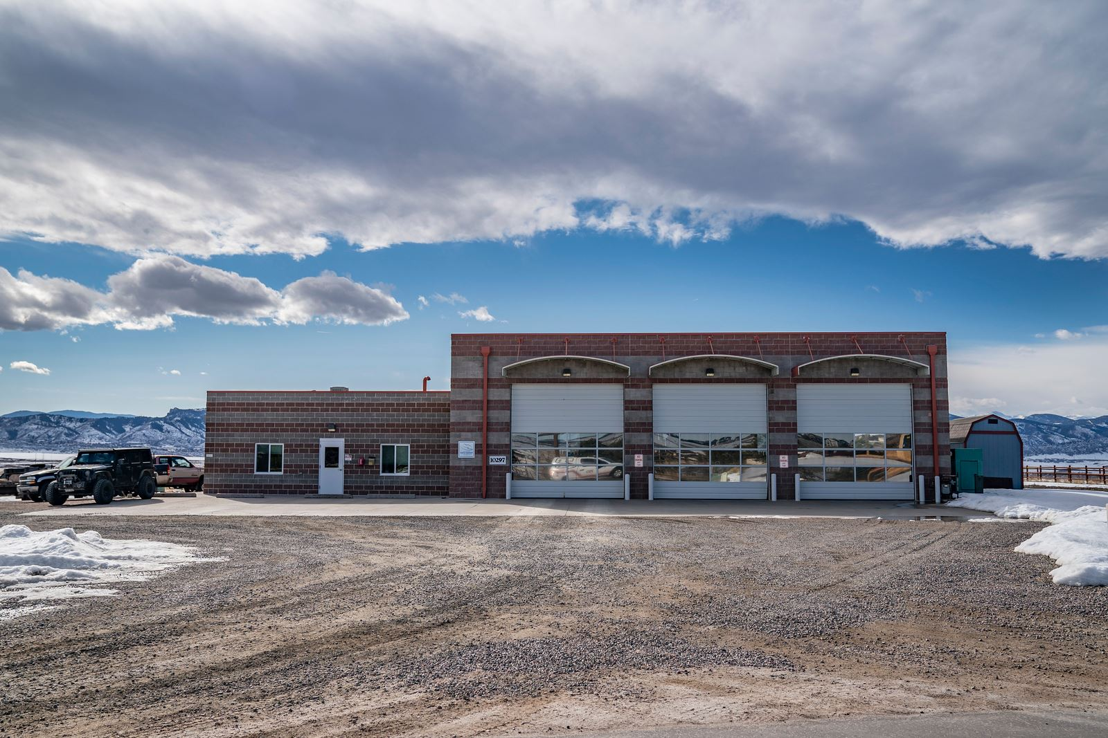 Brick building with garage doors and parked vehicles, set against a mountain backdrop under a cloudy sky.