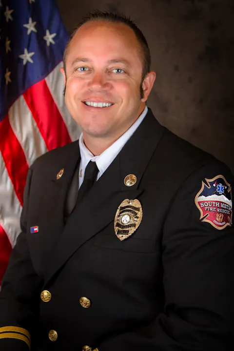 A smiling firefighter in uniform, standing in front of an American flag.