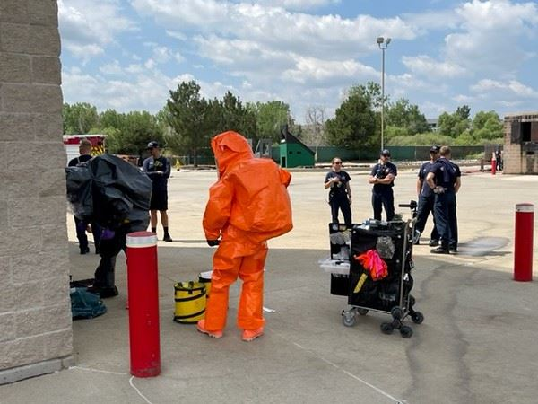 People in a parking lot, one in orange hazmat suit, others in uniform watching; equipment and gear around.